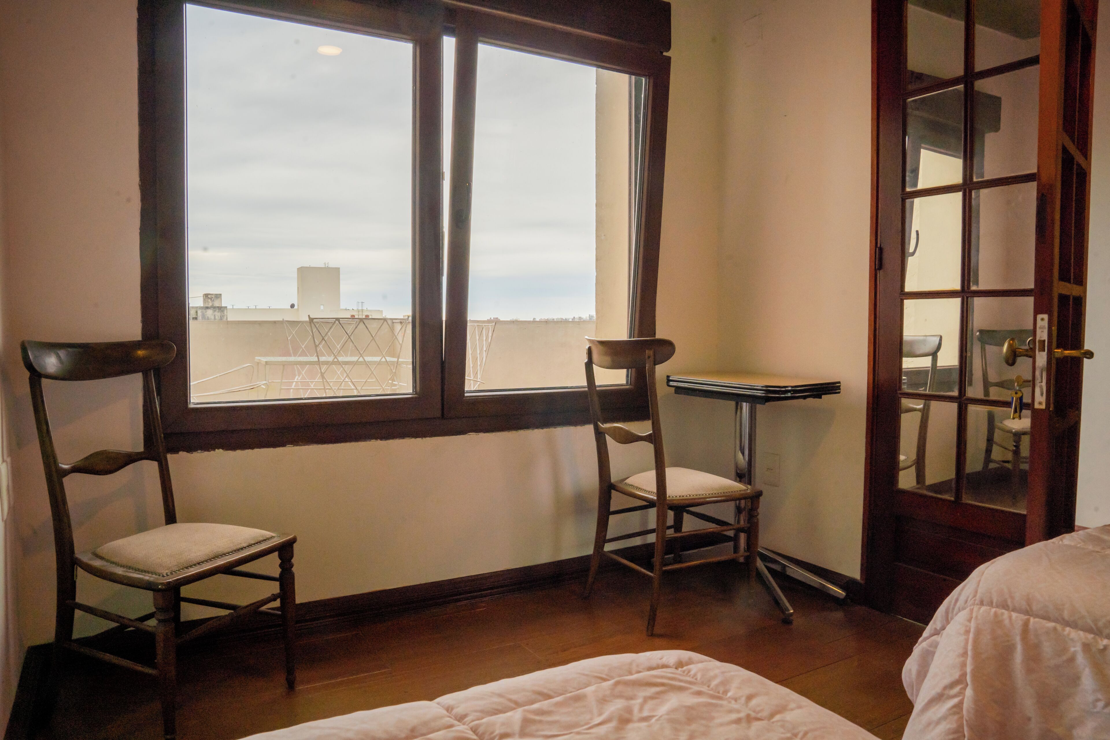 Quarto com duas cadeiras de madeira, uma mesa pequena e uma cama. Há uma janela que oferece vista do céu e de um edifício.