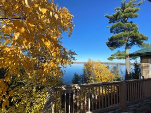 Una terraza de madera con vistas a un lago rodeado de follaje otoñal y un cielo azul despejado.