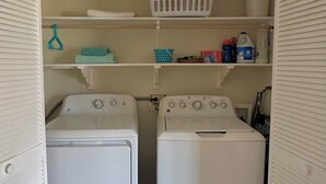 Un lavadero con dos lavadoras blancas de carga frontal, una cesta de ropa blanca y estantes con diversos productos de limpieza.