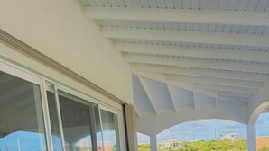 Un balcón techado con un sofá color turquesa y una mesita pequeña, ventanas amplias y vistas a zonas verdes y edificios.