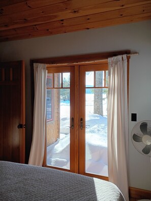 A room with a wooden ceiling, a bed with a quilted cover, a sliding glass door with curtains, and a wall-mounted fan.
