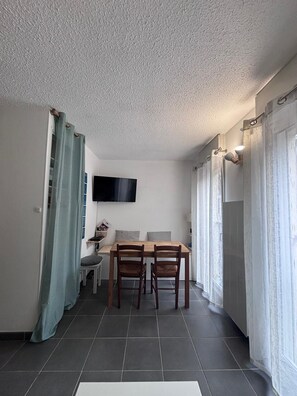 Ein kompakter Wohnbereich mit Esstisch, zwei Stühlen, einem flachbildfernseher an der Wand und einem fenster mit Vorhängen.