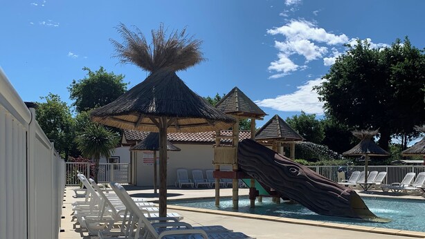 Un área de piscina con sillas de descanso, una palmera y un pabellón con techo de paja.