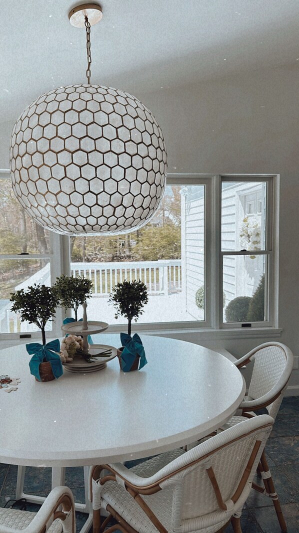 Ett runt matbord med flätade stolar, en hexagonformad hänglampa och två krukväxter dekorerade med blå rosetter.