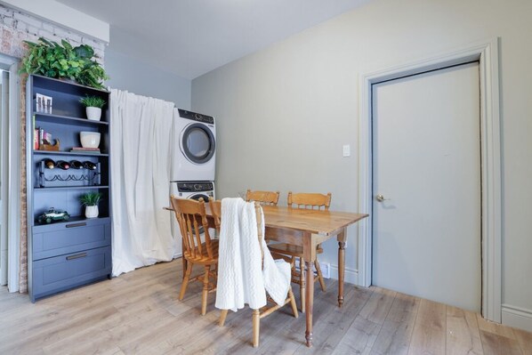 A dining area with a wooden table and chairs, a blue shelving unit with plants, a washing machine, and a door.