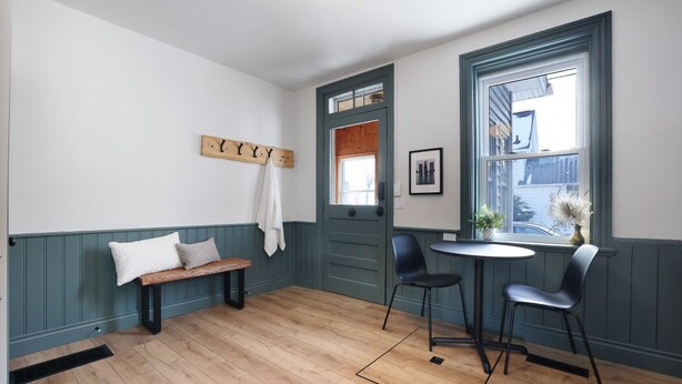 Una habitación con piso de madera, un banco, dos sillas, una mesita, una puerta y una ventana con vistas.