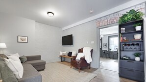 Una sala de estar moderna con un sofá gris, un banco de madera y un sillón de cuero. Hay un televisor de pantalla plana montado en la pared, una estantería con plantas y una pared de ladrillo blanco.