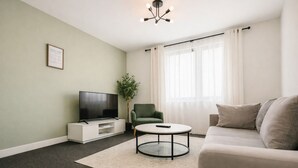 Ein Wohnzimmer mit Flachbildfernseher, einem grünen Sessel, einem runden Couchtisch und einem beigefarbenen Sofa.