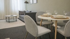 Ein modernes Wohnzimmer mit einem runden Esstisch für zwei Personen, grau gepolsterten Sesseln, einem grauen Sofa, einem kleinen runden Couchtisch und einer Stehlampe.