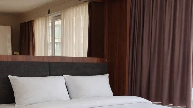 Un llit amb roba de llit blanca en una habitació amb panells de fusta i una finestra gran amb cortines.