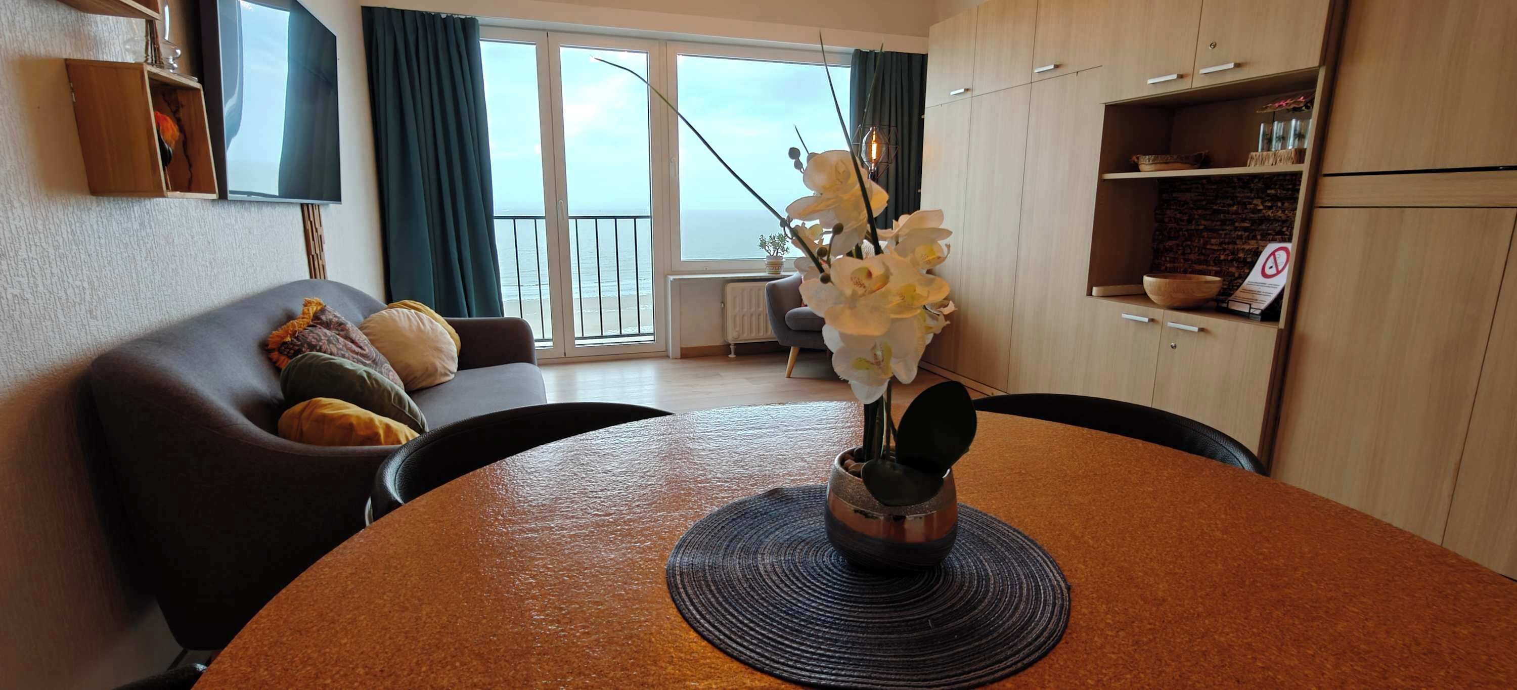 Een woonkamer met een bank, een eettafel met een bloevas, een keuken met kasten, en uitzicht op zee door grote ramen.