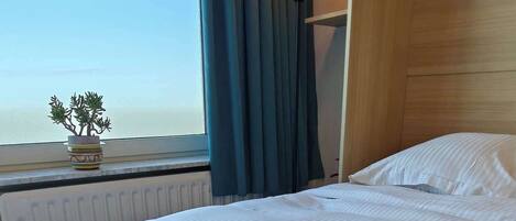 A neatly made bed with folded towels, a wall-mounted lamp, and a view of the sea through a window with curtains.