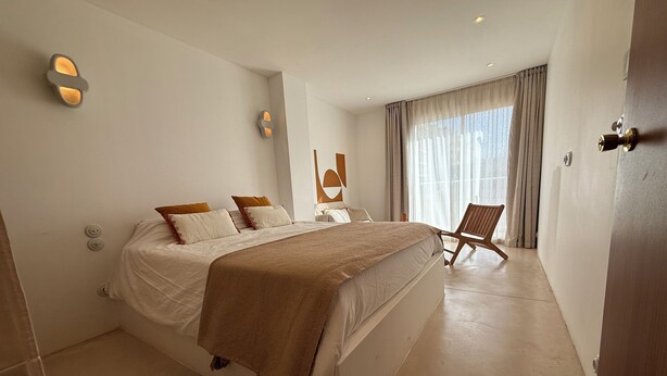 Ein modernes Hotelzimmer mit einem großen Bett, einem Holzsessel und einem großen Fenster mit Vorhängen.