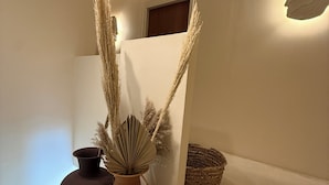Ein Zimmer mit weißer Wand, einer braunen Vase mit Pampasgras, einem dunkelbraunen Topf und einem gewebten Korb.