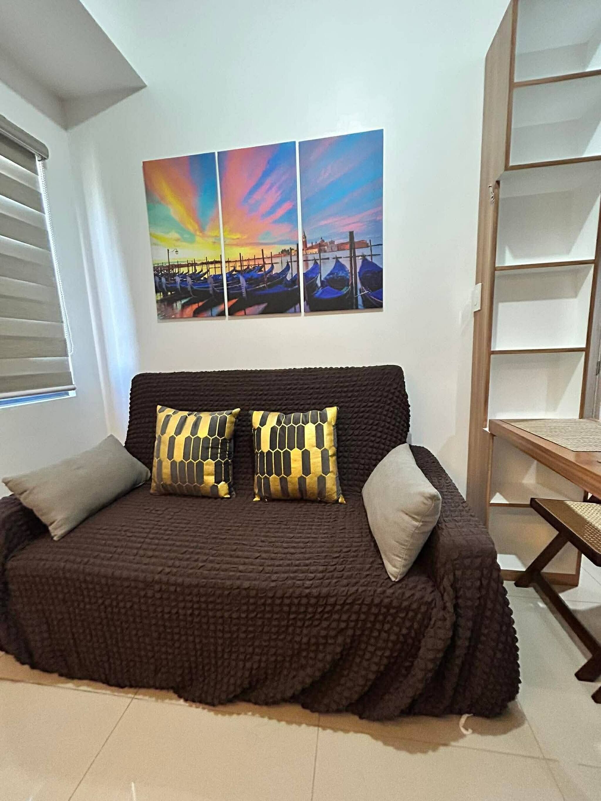 Una sala de estar acogedora con un sofá marrón, dos cojines estampados y un cuadro colgado en la pared que muestra un atardecer sobre un canal.