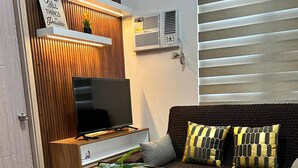 A modern living room with a wooden dining table, wicker chairs, a sofa with patterned cushions, a TV, and a wall-mounted air conditioner. - Lux 2BR Sky Condo With 24-hr Security, Fully Airconditoned & Basement Parking (Dasmarinas)