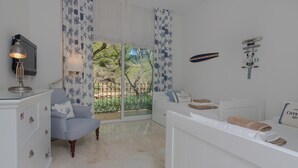 Quarto com cômoda branca, poltrona azul, cama branca e vista para o exterior através de uma porta de correr de vidro.