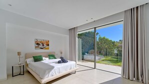 Ein modernes Schlafzimmer mit einem großen Bett, einem Nachttisch, einem Gemälde an der Wand und Blick auf Grün hinter einer Schiebeglastür.
