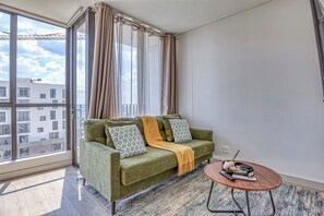 Ein modernes Wohnzimmer mit einem grünen Sofa, gemusterten Kissen, einer gelben Decke und einem kleinen Holztisch mit Fernbedienung und Buch.