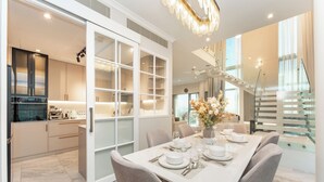A modern dining area with a large table set for six, a kitchen with a built-in oven, and a staircase with glass railings. - PVH - Central Park 1BR with Study Room (Dubai)