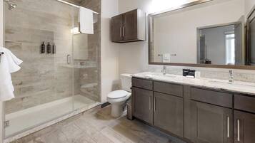 Un baño moderno con cabina de ducha de vidrio, inodoro, lavamanos con un espejo grande y armarios de madera oscura.