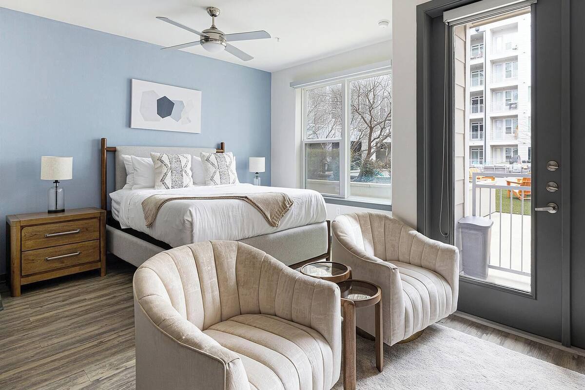 Une chambre à coucher comprenant un lit, des tables de chevet, une chaise, une petite table et une grande fenêtre donnant sur des immeubles.