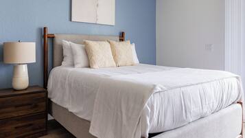 Un lit bien fait, recouvert d’une couette blanche et agrémenté d’oreillers, une table de chevet en bois avec une lampe, et un mur bleu orné d’une œuvre d’art abstraite au-dessus du lit.