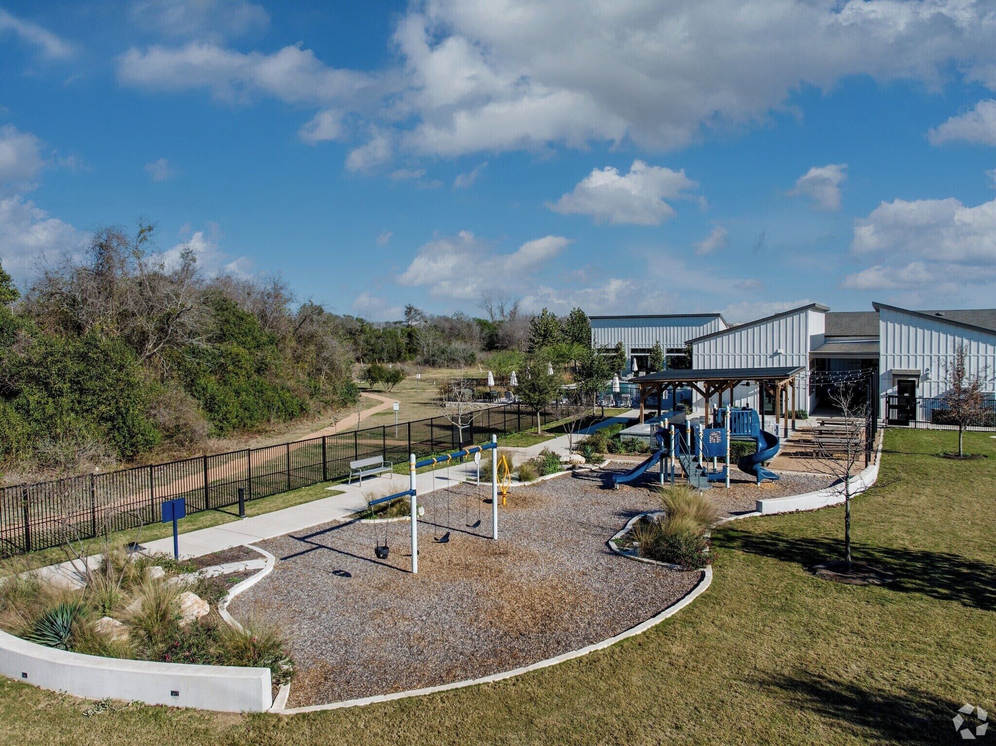 A playground with swings and slides, surrounded by a fence, with a building in the background.