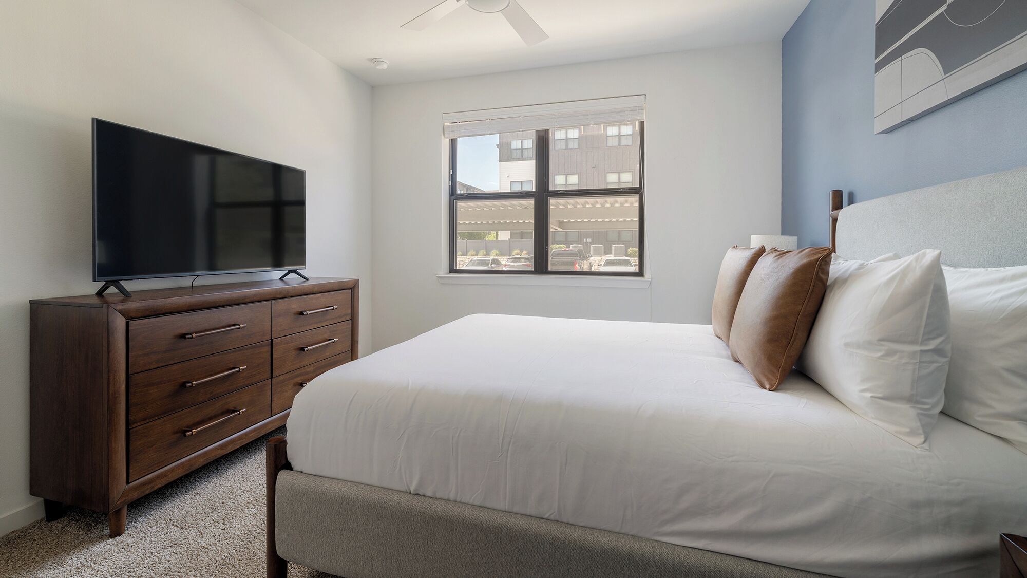 Un dormitorio con cama, un televisor sobre una cómoda de madera, una ventana con vistas a edificios y un ventilador de techo.