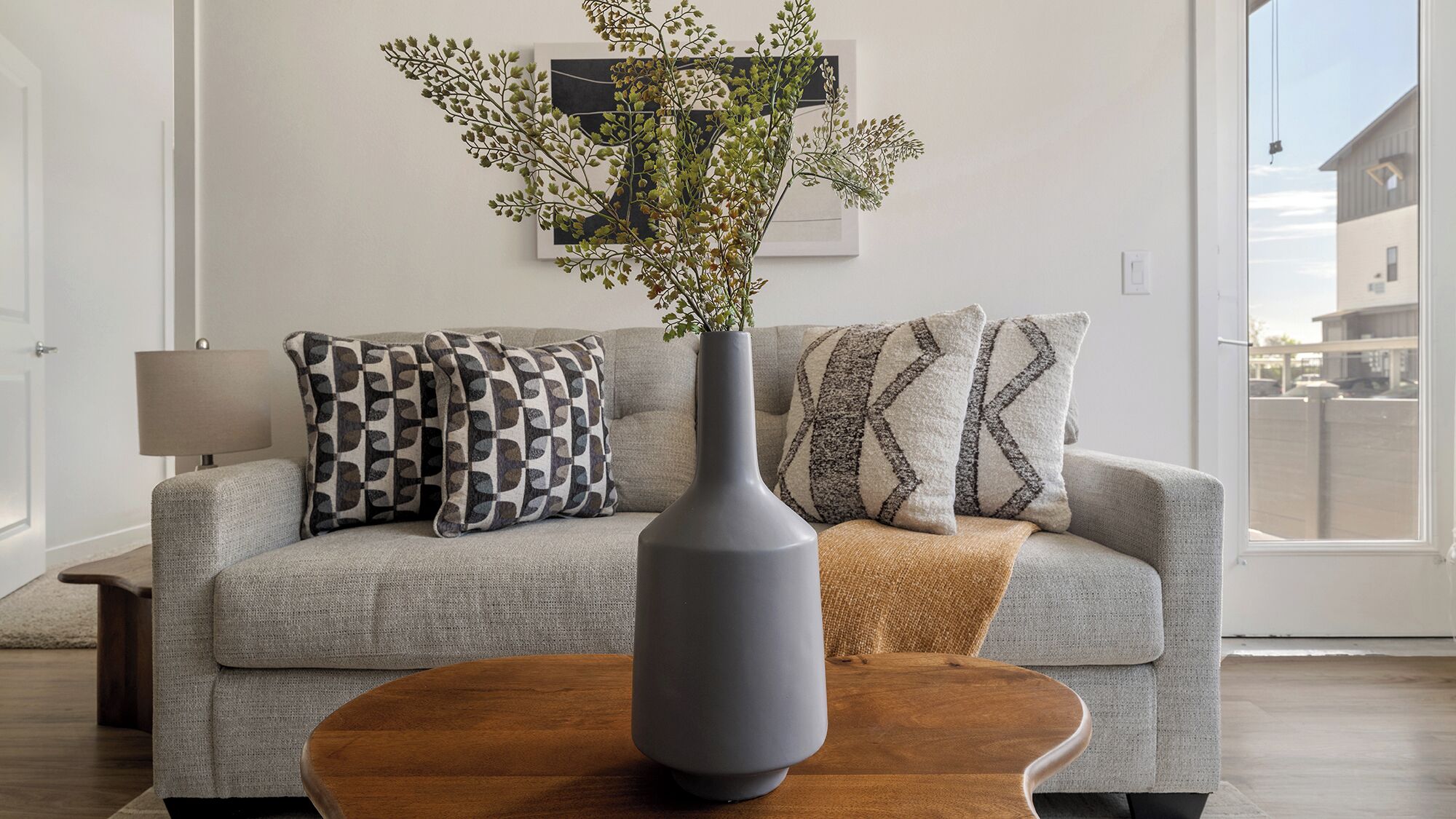 A living room with a grey sofa, patterned cushions, a vase with greenery, and a wooden coffee table.