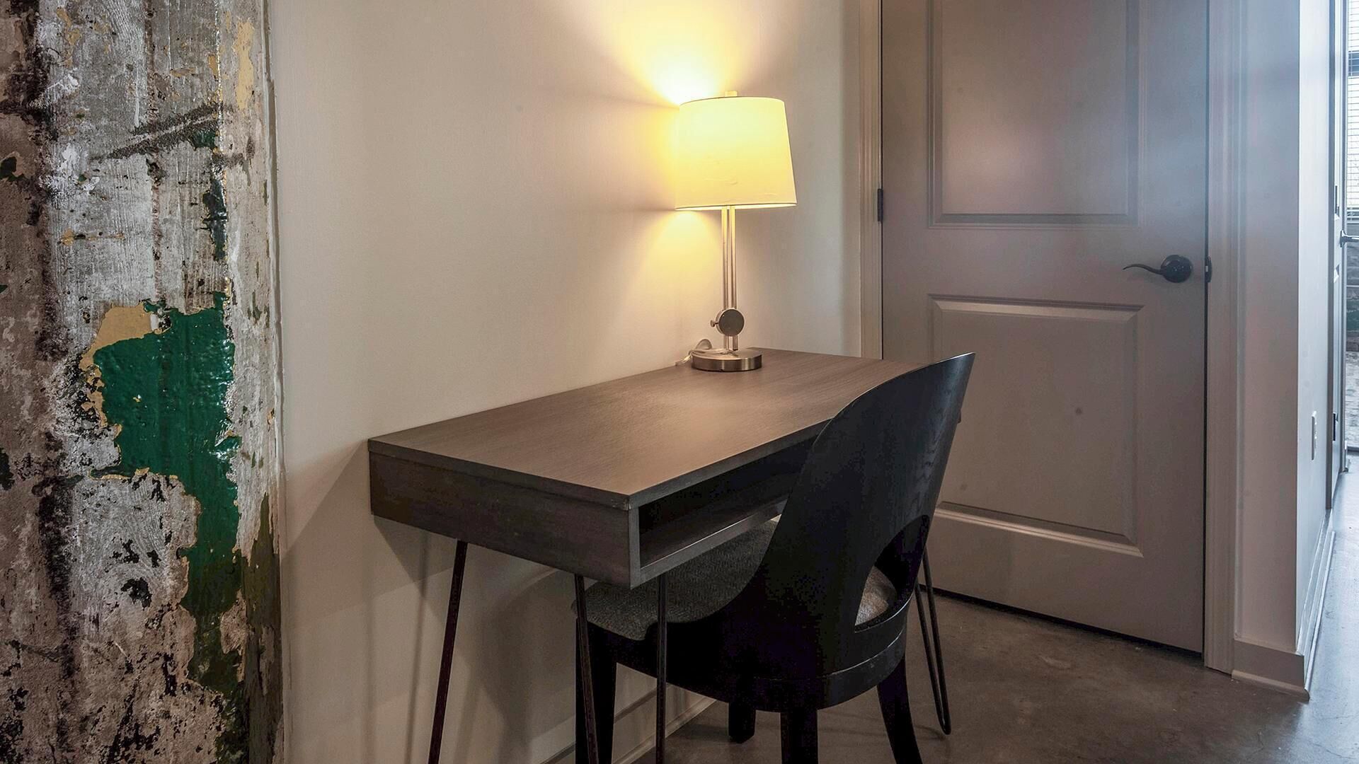 Ein kleiner, dunkler Holztisch mit einer Lampe, ein schwarzer Stuhl und eine weiße Tür in einem Raum mit strukturierter Wand.