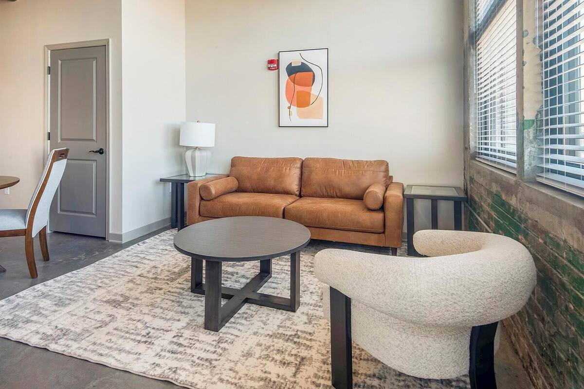 A living room with a brown sofa, a round coffee table, a white armchair, a lamp, and a framed abstract artwork on the wall.
