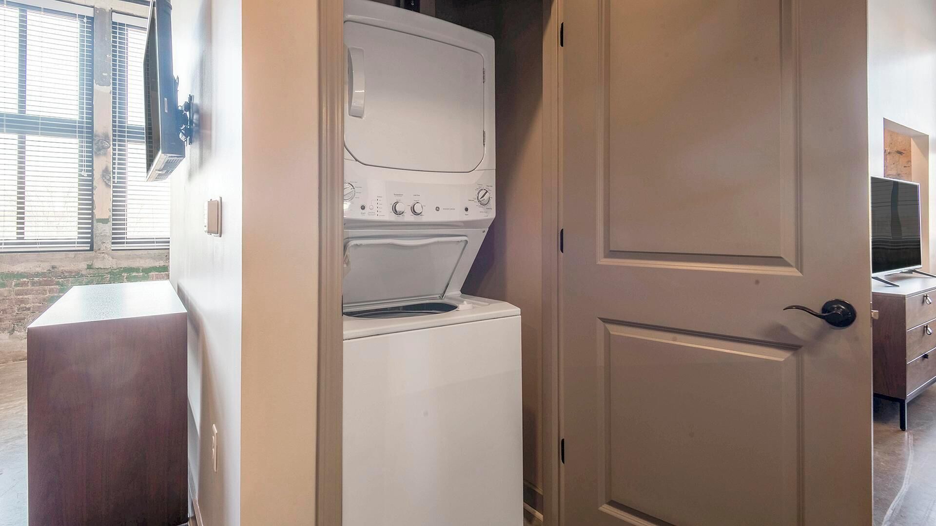 A compact laundry room with a washer and dryer, a wooden bench, and a door leading to another room.