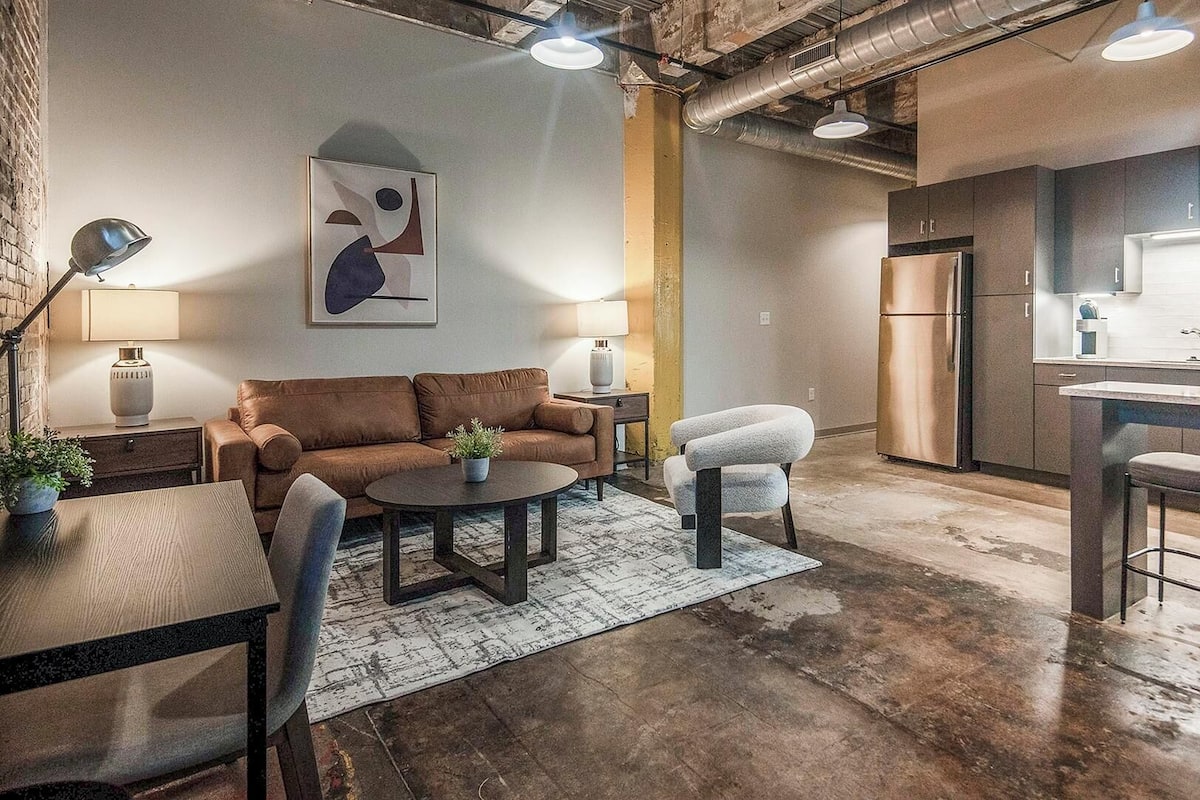 A modern kitchen with a stainless steel refrigerator, a bar area with stools, and a seating area with a sofa and a coffee table.