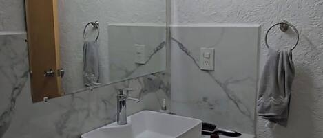 Ein Badezimmer mit weißem Waschbecken, Spiegel und Marmorfliesen. Handtuchhalter, Mülleimer und Toilette sind vorhanden.