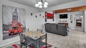 Un comedor con mesa y sillas de vidrio, un cuadro de un autobús de dos pisos rojo, un sofá gris y un televisor montado en la pared.