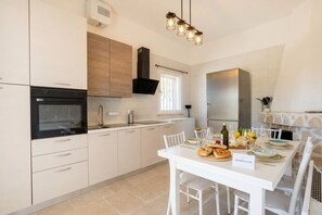Une cuisine moderne avec des meubles blancs, un four encastré, une hotte en acier inoxydable et un coin repas avec une table blanche mise pour deux personnes.