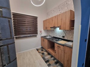 Une cuisine de taille réduite, dotée de meubles en bois, d’une cuisinière à gaz et d’une fenêtre avec des stores.