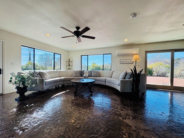 A spacious living room with a curved sectional sofa, a coffee table, and a ceiling fan. Large windows provide views of the outdoors.