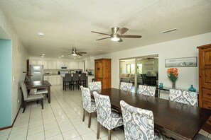 A spacious dining area with a large wooden table and patterned chairs, a kitchen with white cabinets, and ceiling fans.