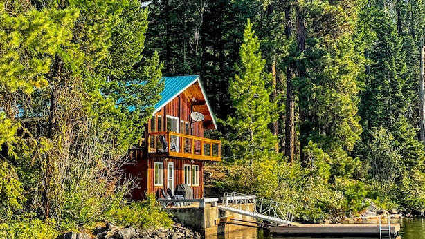 Uma cabana com telhado azul, cercada por uma floresta densa, perto de um píer que se estende sobre um lago.
