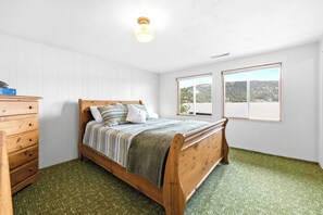 A bedroom with a wooden bed, a dresser, and two windows offering a view of a forested landscape. - Secluded Lakeside Cabin, Dock, Deck, Lake Views (McCall)