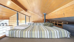 Quarto com teto de madeira, cama com roupa de cama listrada, janela e uma pequena mesa com recipientes.