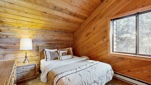 Une chambre à coucher dans un chalet en bois, avec un lit, une table de chevet, une lampe et une fenêtre donnant sur la forêt.