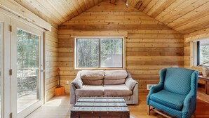 L’intérieur d’un chalet accueillant, avec un plafond en bois, un canapé confortable, un fauteuil bleu et une grande fenêtre donnant sur la forêt.