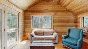 Ein gemütliches Blockhaus-Interieur mit Holzbalkendecke, einer gepolsterten Couch, einem blauen Sessel und einem großen Fenster mit Blick auf Bäume.
