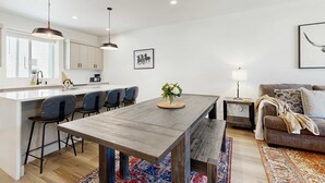A modern kitchen with white cabinets, a central island with bar stools, pendant lighting, and a dining table with a vase of flowers. - Modern Townhouse, Centrally Located Near BSU (Boise)