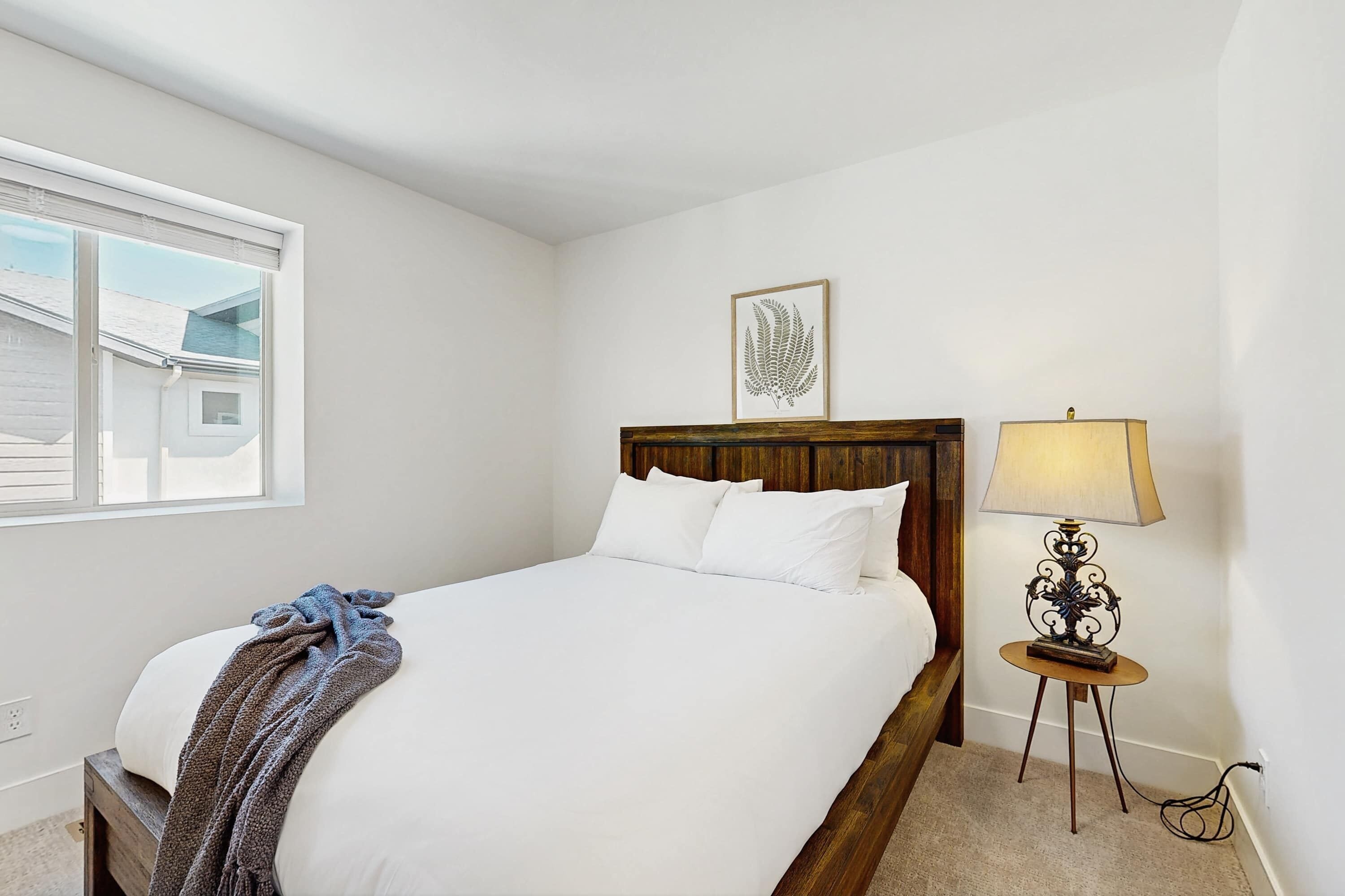 A bedroom with a bed, a bedside lamp, a framed artwork, and a window.