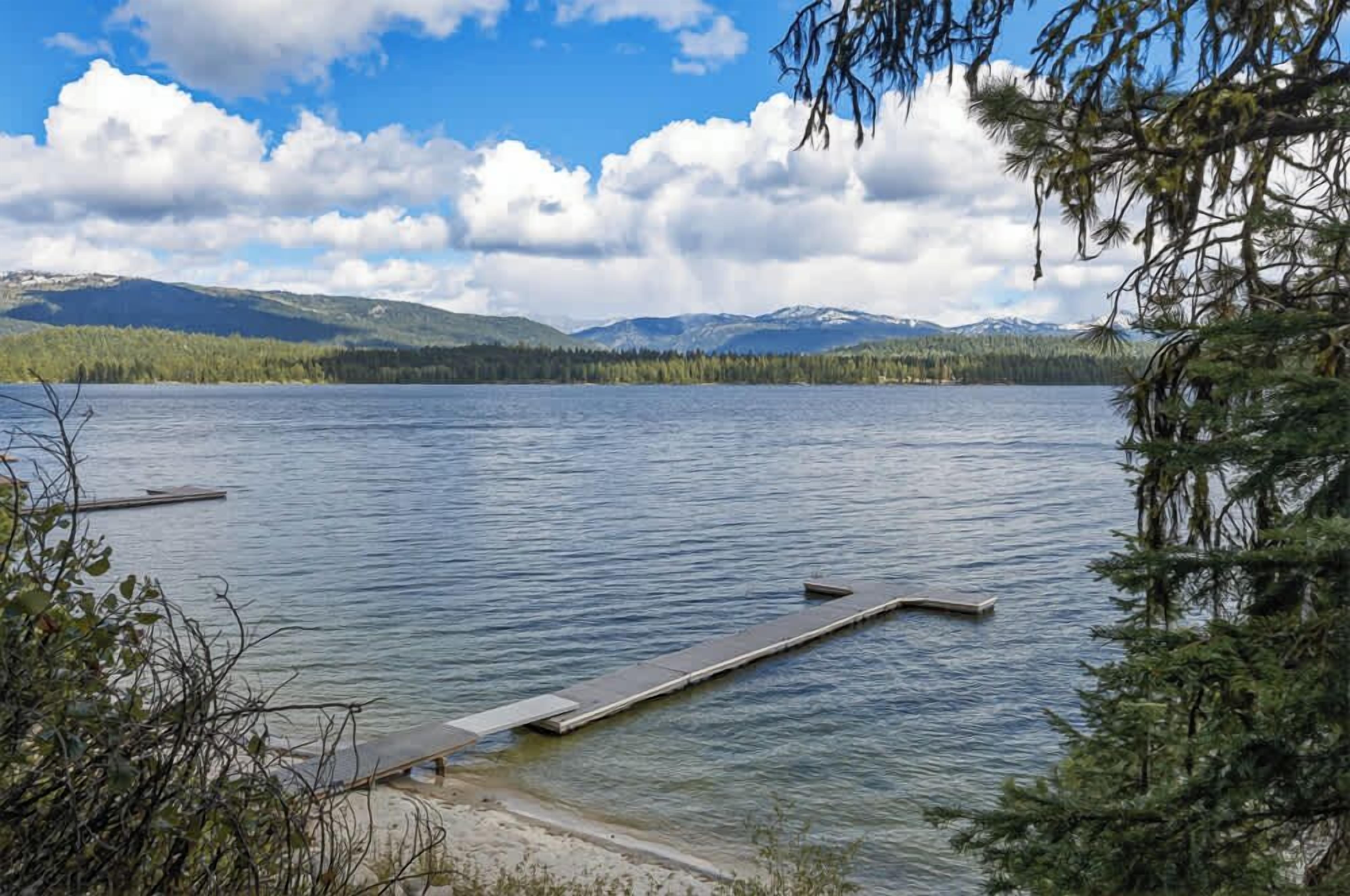A calm lake with a dock extending into the water, surrounded by trees and mountains in the background.