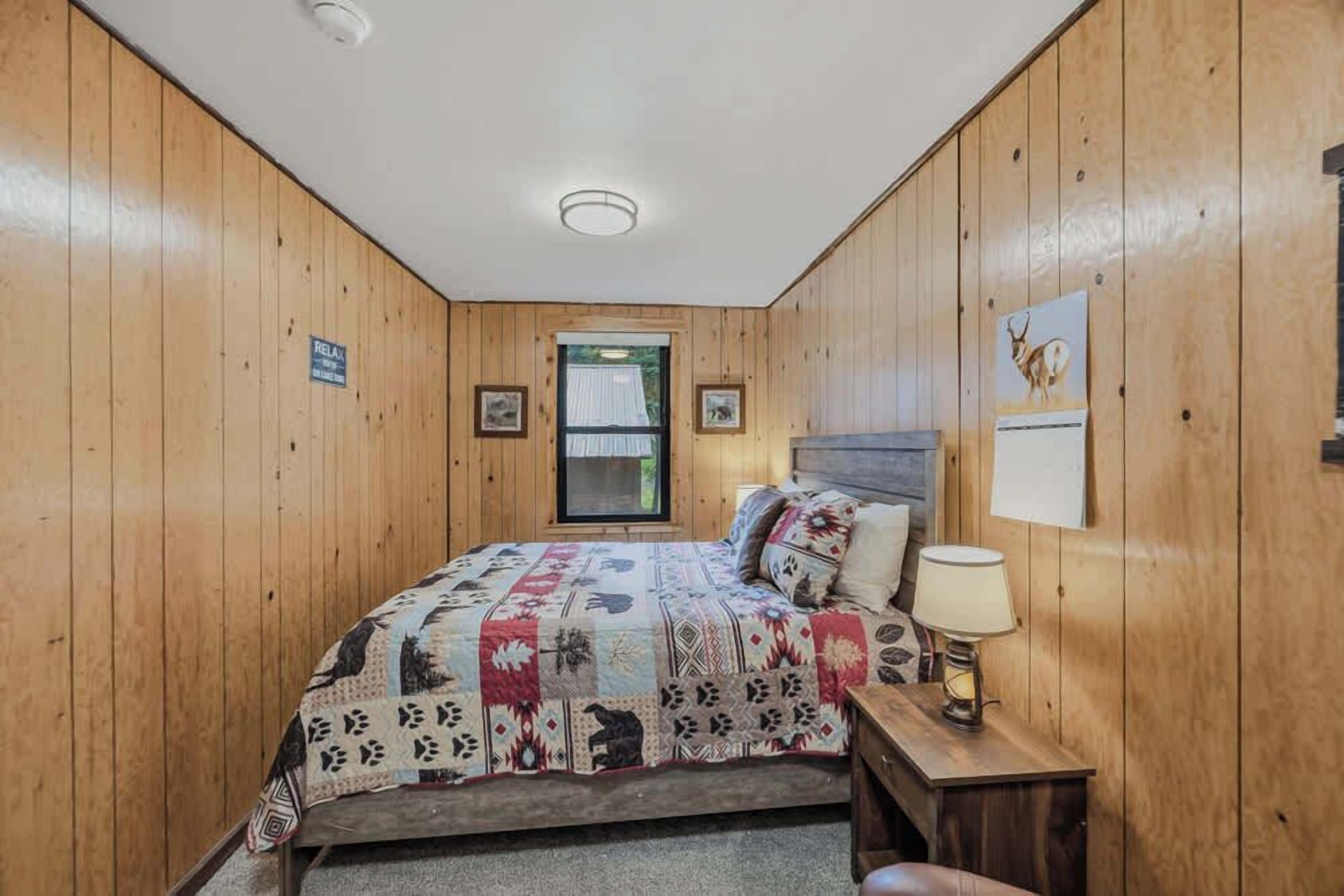 A wooden-paneled room with a bed, a nightstand, a lamp, and framed pictures on the wall.